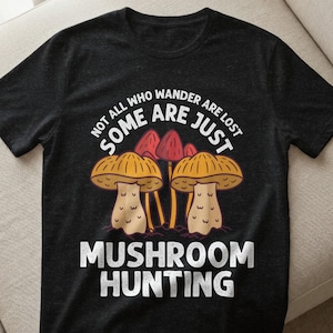 Könnte beinhalten: Dunkelgraues T-Shirt mit dem Thema Pilzsuche. Das Shirt zeigt eine Grafik von mehreren Pilzen in Gelb-, Rot- und Brauntönen. Der Text auf dem Shirt lautet: "Not all who wander are lost, some are just mushroom hunting."