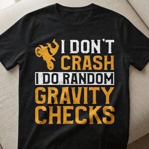 Könnte beinhalten: Schwarzes T-Shirt mit dem Text "I DON'T CRASH I DO RANDOM GRAVITY CHECKS" in Weiß und Orange. Auf der linken Seite befindet sich eine Motorradgrafik. Das T-Shirt ist aus weichem Material.