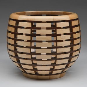 May include: A handcrafted wooden basket with a unique woven design. Alternating light and dark wood strips create a geometric pattern. The basket has a rounded shape with a flat base and a wide opening, perfect for decorative storage.