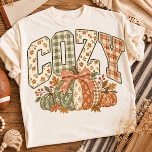 Op de afbeelding: Een crèmekleurig t-shirt met het woord COZY in grote blokletters met diverse patronen, waaronder groen ruitpatroon, bloemenprints en oranje ruitpatroon. Onder de tekst staan drie pompoenen versierd met bloemmotieven en een strik.