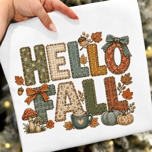 May include: A white t-shirt featuring the text HELLO FALL in large, patterned block letters. The design includes autumn-themed illustrations such as mushrooms, a mug of hot cocoa, pumpkins, acorns, and leaves in shades of green, orange, and brown.