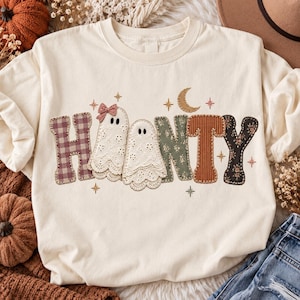 Könnte beinhalten: Ein cremefarbenes T-Shirt mit dem Wort "HAUNTY" in großen, strukturierten Buchstaben. Das "H" ist lila kariert, das "A" und "U" sind weiße Spitzengeister, das "N" ist grün geblümt, das "T" ist brauner Kord und das "Y" ist schwarz geblümt.