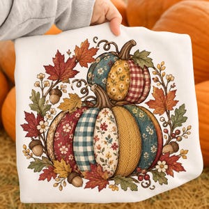 May include: A white sweatshirt featuring a graphic of two stacked pumpkins decorated with various patterns including floral prints, gingham, and plaid. The pumpkins are surrounded by a wreath of autumn maple leaves, oak leaves, acorns, and small white flowers.