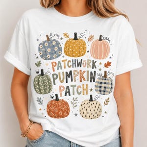 May include: A white short-sleeve t-shirt featuring a graphic of seven pumpkins with various patterns including floral, polka dot, and plaid. The text PATCHWORK PUMPKIN PATCH is printed in the center in multi-colored block letters surrounded by small stars and leaves.