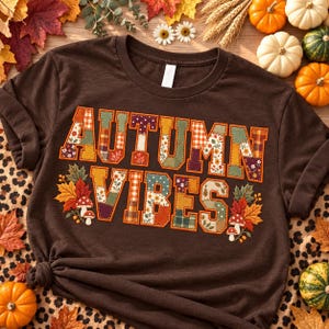 May include: A dark brown t-shirt featuring the text AUTUMN VIBES in a patchwork-style font with various patterns including plaid, floral, and polka dots. The design is accented with illustrations of colourful autumn leaves and small mushrooms.