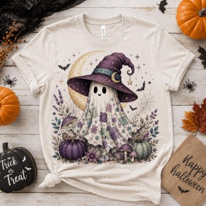 Könnte beinhalten: Ein cremefarbenes T-Shirt mit einer Grafik eines Geistes, der einen violetten Hexenhut trägt. Der Geist ist mit violetten und blaugrünen Blumenmustern verziert und von einer Mondsichel, Fledermäusen, Spinnenweben, Pilzen sowie violetten und grünen Kürbissen umgeben.