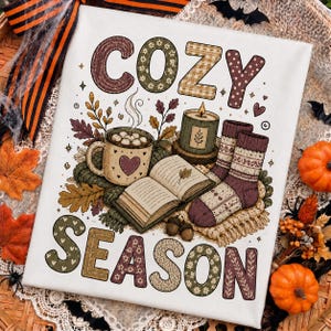 May include: A white canvas print featuring the text COZY SEASON in patterned block letters. The illustration includes a steaming mug with a heart, an open book with a leaf, a lit candle, and a pair of knit socks, all surrounded by autumn leaves.