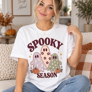 May include: A white short-sleeved t-shirt featuring a graphic of three ghosts with floral, moon, and star patterns. The text SPOOKY SEASON is printed in dark red above and below the ghosts, which are accompanied by small orange pumpkins.