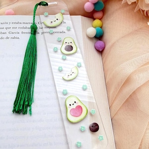 May include: A clear acrylic bookmark featuring avocado and melon illustrations with smiling faces and a green tassel. The bookmark is decorated with small blue flowers and a brown dot. The bookmark is placed on top of an open book.