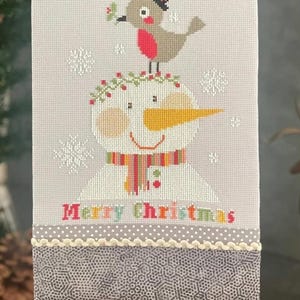 May include: A stitched Christmas decoration featuring a snowman with a bird wearing a top hat. The snowman has a carrot nose and a colorful scarf. The words "Merry Christmas" are stitched at the bottom.