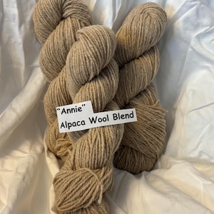 Op de afbeelding: Twee strengen "Annie" Alpaca Wool Blend garen in een natuurlijke beige kleur. Het garen is dik en getwist, met een zacht, getextureerd uiterlijk. De strengen worden gepresenteerd op een witte stoffen achtergrond.