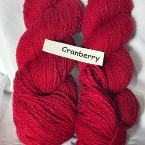 May include: Four skeins of deep red yarn, labelled "Cranberry". The yarn is tightly wound, with a textured surface. The colour is a rich, vibrant red, suitable for knitting or crochet projects. The yarn is displayed against a white background.