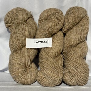 May include: Three skeins of oatmeal-coloured yarn are shown. A white label with the word "Oatmeal" is placed in front of the yarn. The yarn appears to be a natural fibre, suitable for knitting or crocheting projects.