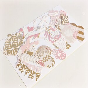 May include: A collection of pink and gold paper cutouts with various geometric and floral designs. The cutouts are arranged on a white background.