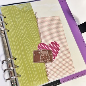 May include: A purple planner with a green and white patterned page insert. The insert features a brown camera with a heart in the lens and a pink heart with white polka dots.