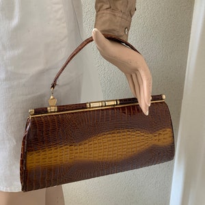 Peut inclure: Un sac à main marron vintage avec un motif peau de crocodile. Le sac à main a une forme rectangulaire avec un fermoir et une poignée dorés. Le sac est tenu par une main de mannequin.