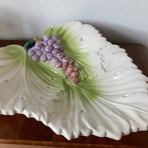 Könnte beinhalten: Eine weiße Keramik-Servierplatte in Form eines grünen Blattes mit einem erhabenen Design aus Trauben. Die Trauben sind lila und rosa.