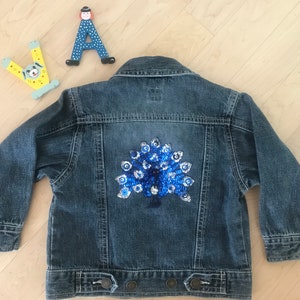 Könnte beinhalten: Eine blaue Jeansjacke mit einem Pailletten-Pfauenmuster auf der Rückseite.