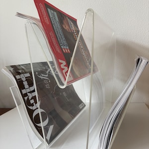 May include: A clear acrylic magazine rack with a modern design. The rack holds several magazines, including one with a red cover and the word "NEW" visible. Another magazine has the word "VOGUE" on the cover. The rack is on a white surface.