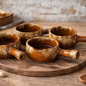 May include: Four brown ceramic soup bowls with handles, displayed on a wooden serving board. The bowls have a rustic, glazed finish with brown and cream tones. A wooden spoon and basket of bread are in the background.