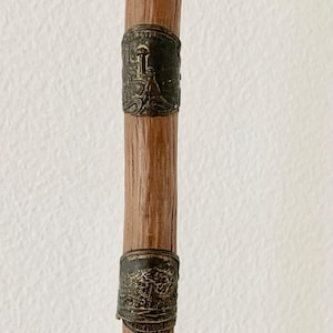 May include: A wooden walking stick with decorative metal bands. The bands are dark with detailed relief designs, possibly depicting historical scenes or landmarks. The wood has a natural, light brown color.