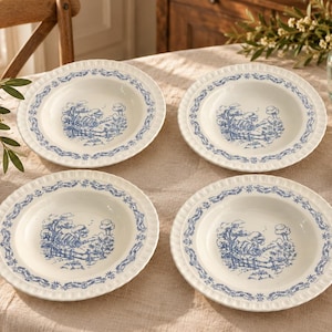 May include: A set of four round ceramic soup bowls featuring a cream-colored base with a blue transferware pattern. Each bowl displays a central landscape illustration of a house and trees, surrounded by a decorative floral border on the rim.