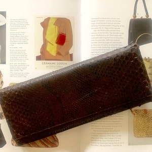 Op de afbeelding: Een bruine leren clutch met een textuur. De clutch ligt op een pagina van een boek over mode.