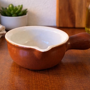 Peut inclure: Un bol à soupe en céramique marron avec un intérieur blanc et une poignée. Le bol a un bec verseur. La poignée est arrondie et lisse. Le bol est posé sur une surface en bois.