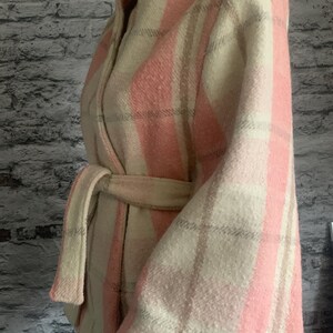 Puede incluir: Una chaqueta cruzada de manga larga con un patr&oacute;n de cuadros texturizado en tonos rosa claro, crema y beige. La prenda incluye un cintur&oacute;n de tela a juego en la cintura, un cuello ancho y costuras blancas visibles a lo largo del dobladillo.