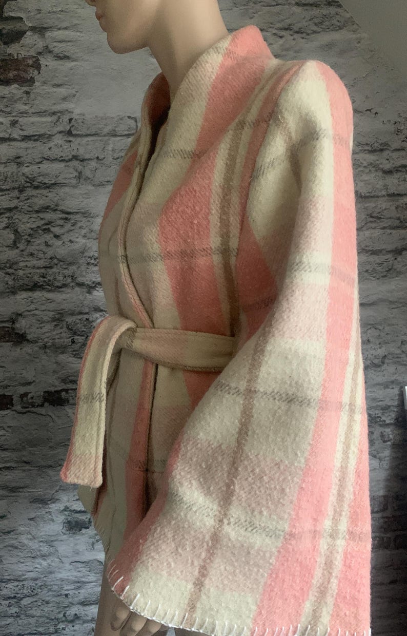 Puede incluir: Una chaqueta cruzada de manga larga con un patr&oacute;n de cuadros texturizado en tonos rosa claro, crema y beige. La prenda incluye un cintur&oacute;n de tela a juego en la cintura, un cuello ancho y costuras blancas visibles a lo largo del dobladillo.