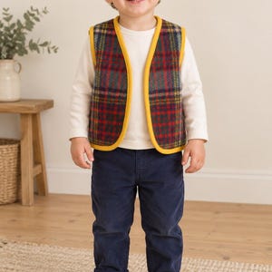 Puede incluir: Un chaleco sin mangas con estampado de cuadros en rojo, verde y amarillo con ribete amarillo sólido, usado sobre una camiseta de manga larga color crema, combinado con pantalones de pana azul oscuro y zapatillas marrón claro con velcro.