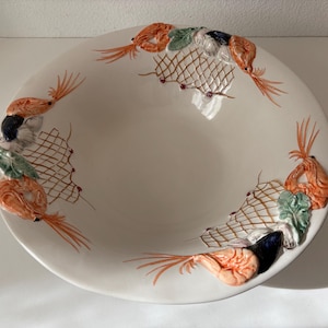 Puede incluir: Un gran cuenco de cerámica blanquecina con un diseño de marisco. El borde está decorado con gambas naranjas, algas verdes y redes de pesca marrones. El cuenco es redondo y tiene una forma ancha y poco profunda.