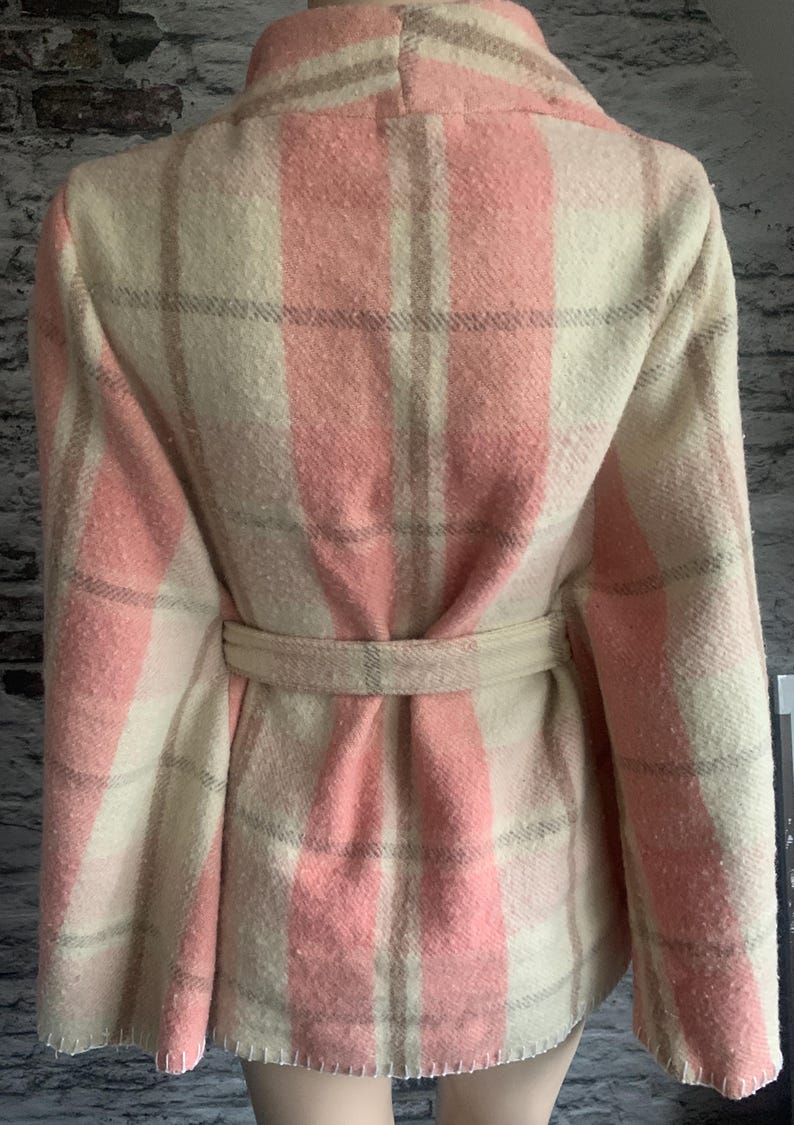 Puede incluir: Una chaqueta de manga larga con un patr&oacute;n de cuadros en tonos rosa suave, crema y gris claro. La prenda incluye un cintur&oacute;n de tela a juego en la cintura, un cuello ancho y costuras visibles a lo largo del dobladillo inferior texturizado.