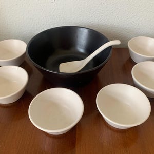 Puede incluir: Un juego de siete cuencos blancos, incluyendo un cuenco grande negro con una cuchara de servir blanca. Los cuencos están dispuestos sobre una superficie de madera.
