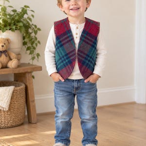 Puede incluir: Un chaleco sin mangas con un patrón de cuadros en rojo, azul y verde con costuras visibles en los bordes. El chaleco se lleva sobre una camisa henley de manga larga de color crema con botones, combinada con vaqueros azules y zapatillas gris claro.