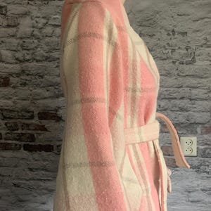 Puede incluir: Un abrigo de manga larga con un patr&oacute;n de cuadros a gran escala en tonos rosa suave y crema. La prenda incluye un cuello ancho, un cintur&oacute;n de tela a juego en la cintura y costuras visibles a lo largo del dobladillo inferior.