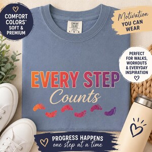May include: A blue crew neck t-shirt featuring the text EVERY STEP Counts in a gradient of orange, red, and purple, accompanied by a row of colourful footprint graphics. The shirt has a Comfort Colors label at the neck and a size L-G-G tag.