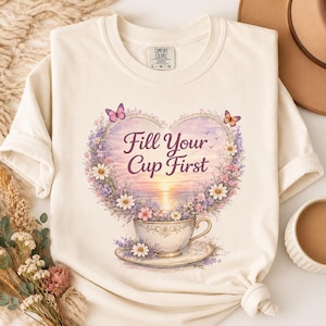 Könnte beinhalten: Ein cremefarbenes T-Shirt mit einer Grafik einer Teetasse, die mit einem herzförmigen Blumenarrangement und Schmetterlingen gefüllt ist. Der Text "Fill Your Cup First" steht mittig im Herzen, das einen Sonnenuntergang über dem Meer einrahmt.