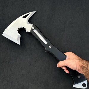 May include: A tactical axe with a silver blade and a black handle. The axe head features a sharp blade, a curved spike, and a gear-shaped cutout. The handle has a streamlined design with a white accent.