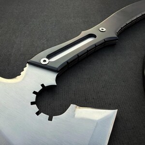 May include: A close-up of a large, silver and black blade. The blade has a unique shape with a serrated edge and a circular cutout. The handle is black with silver rivets. The blade is set against a dark background.