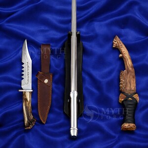 May include: An assortment of blades and knives are displayed on a blue satin surface. The collection includes a long, slender silver blade in a black stand, a knife with a serrated edge and antler handle, and a curved knife with a wooden handle.