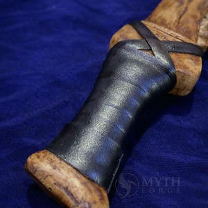 May include: Close-up of a handcrafted knife handle. The handle is made of light-colored wood with a dark leather grip. The leather is wrapped around the handle and secured with a leather strap. The knife handle is set against a blue background.