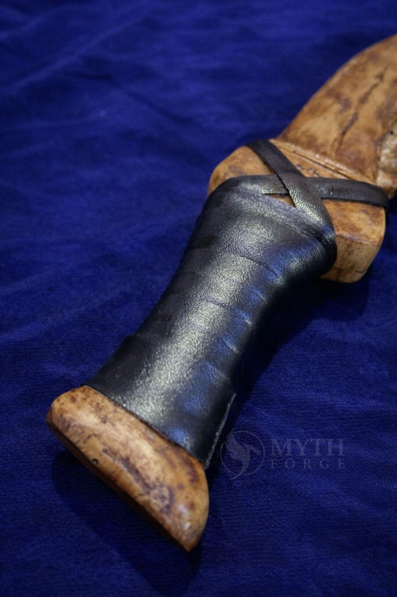 May include: Close-up of a handcrafted knife handle. The handle is made of light-colored wood with a dark leather grip. The leather is wrapped around the handle and secured with a leather strap. The knife handle is set against a blue background.