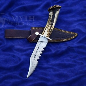 May include: A hunting knife with a serrated blade and a handle made from an antler. The knife rests in a brown leather sheath with gold stitching, secured by a leather strap. The knife has etched text on the blade.