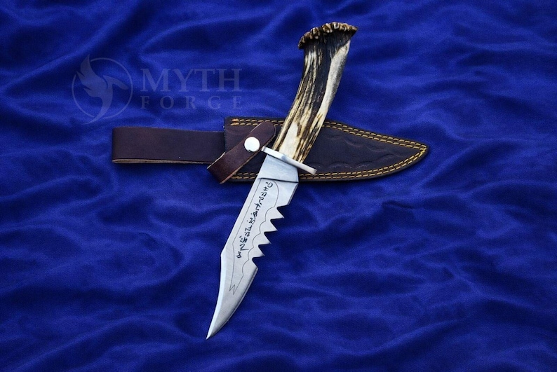 May include: A hunting knife with a serrated blade and a handle made from an antler. The knife rests in a brown leather sheath with gold stitching, secured by a leather strap. The knife has etched text on the blade.