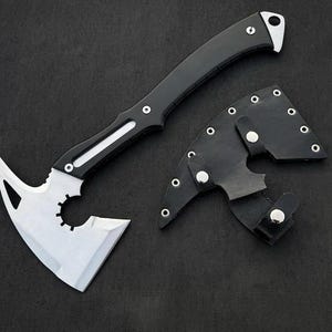 May include: A tactical axe with a black handle and a silver blade. The blade has a unique cutout design. A black leather sheath with silver rivets is included. The axe is designed for outdoor use.