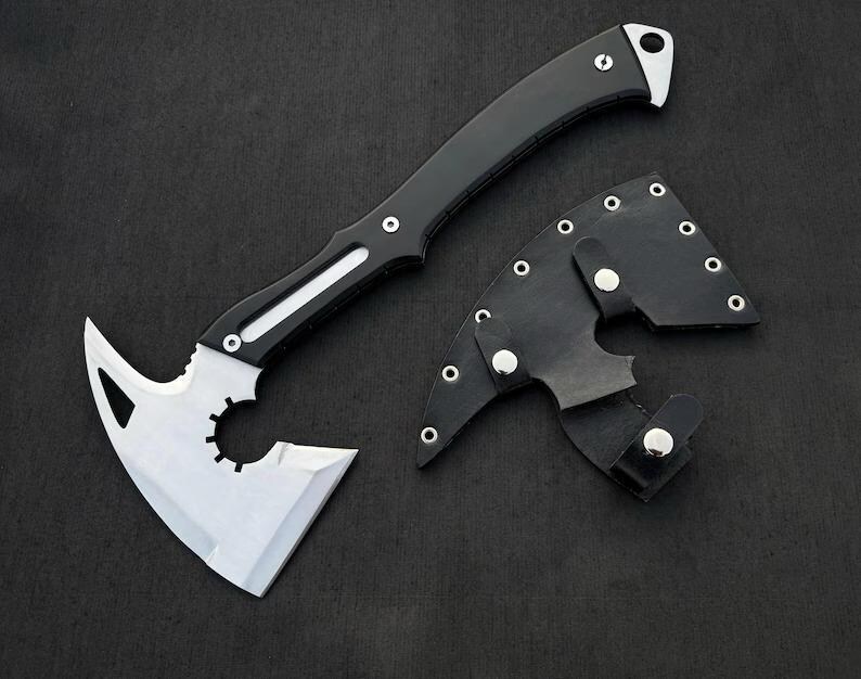 May include: A tactical axe with a black handle and a silver blade. The blade has a unique cutout design. A black leather sheath with silver rivets is included. The axe is designed for outdoor use.