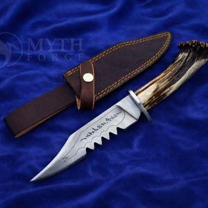 May include: A hunting knife with a serrated blade and a handle made from an antler. The knife is accompanied by a brown leather sheath. The blade has an inscription. The knife and sheath are on a blue surface.