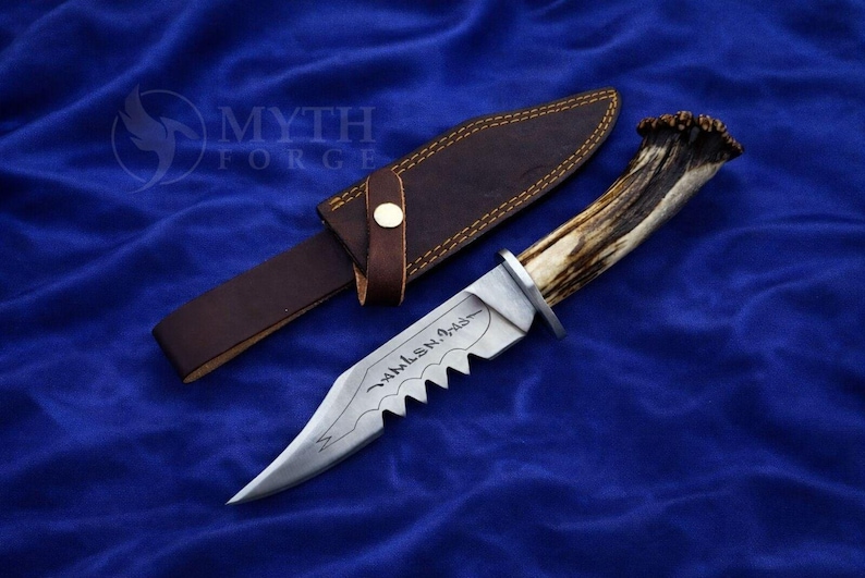 May include: A hunting knife with a serrated blade and a handle made from an antler. The knife is accompanied by a brown leather sheath. The blade has an inscription. The knife and sheath are on a blue surface.