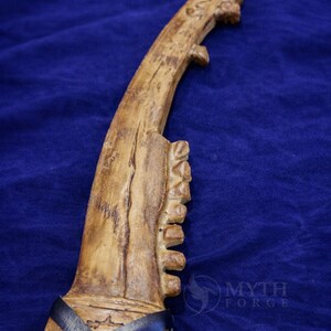 May include: A carved, bone-colored dagger with a curved blade and a textured handle. The handle is wrapped with black leather straps. The dagger has a unique, rustic appearance, suggesting a handmade or ancient origin. The Myth Forge logo is visible.